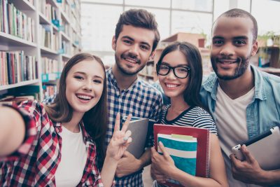 Selfie time! Four students with beaming smiles are posing for selfie shot, caucasian attractive lady is taking, in school library building. Gathered, cheerful, smart and successful youth