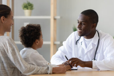 Smiling black male doctor listen cute african boy patient tell complaint make notes at medical consultation, mom and child son visit African pediatrician talk in clinic, children healthcare concept